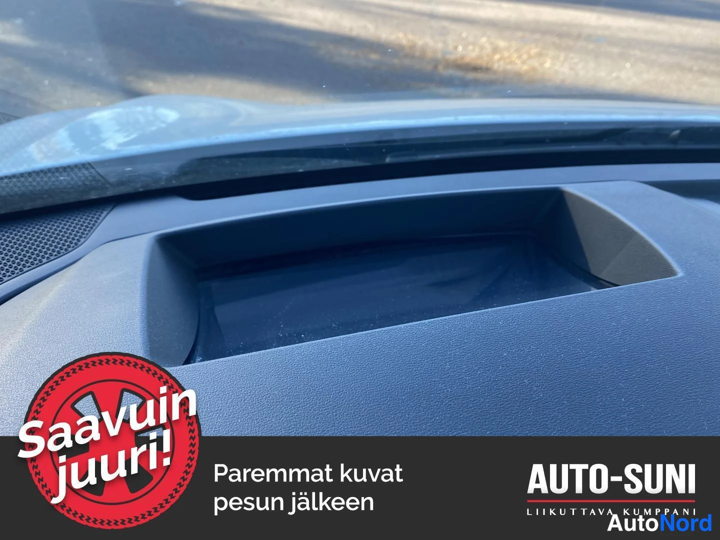 Muu: Kojelaudan yläosan lähikuva: tummanharmaassa panelissa syvennetty näyttöaukko ja vasemmalla kaiuttimen ritilä, tuulilasin takana sumeneva katu. Kuvan alaosassa