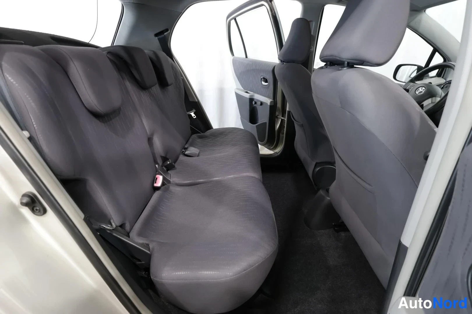 Muu: Sisätilakuva: takaistuimen selkänojat ja etuistuinten selkänojat harmaassa kangasverhoilussa, avattu takaovi ja ohjauspyörä Toyota-tunnuksella näkyvät.