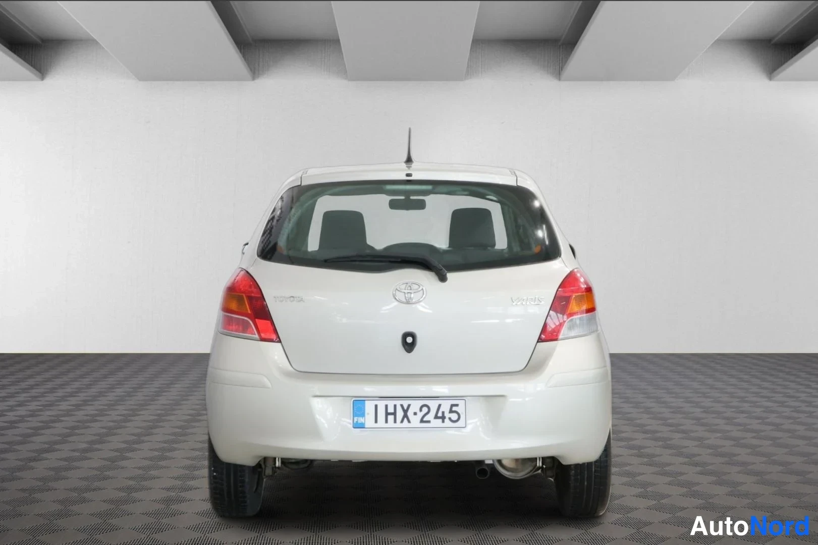 Takaa: Toyota Yaris -henkilöauto suoraan takaa; vaaleanbeige kori, takavalojen väissä Toyota-merkki sekä TOYOTA- ja YARIS-tekstit, takilahinpesuri, rekisteritunnus ja