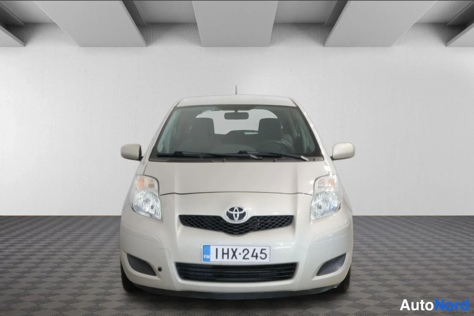 Edestä: Vaaleanbeige Toyota Yaris -henkilöauto kuvattuna suoraan edestä, rekisterikilpi.