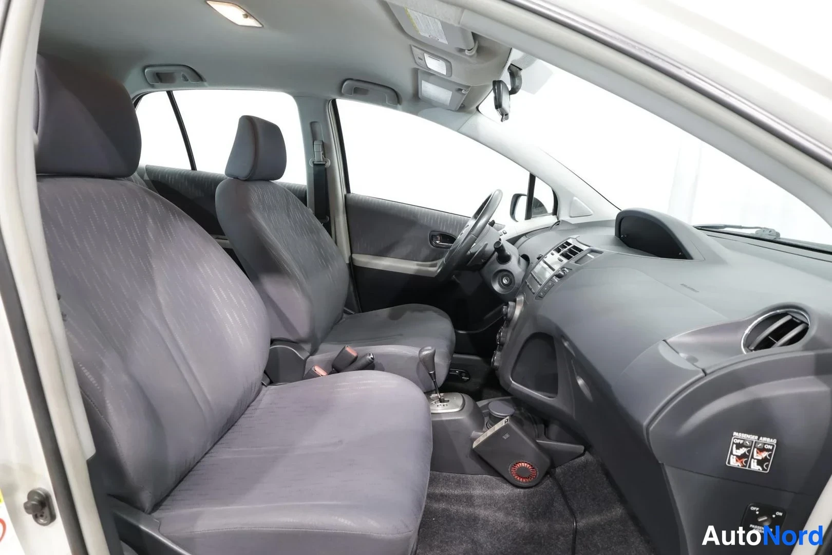 Etuviisto, nokka oikealle: Toyota Yarisin sisätilat avatusta ovesta kuvattuna: kaksi tummaa etuistuinta, ohjauspyörä, vaihteenvalitsin ja kojelauta.