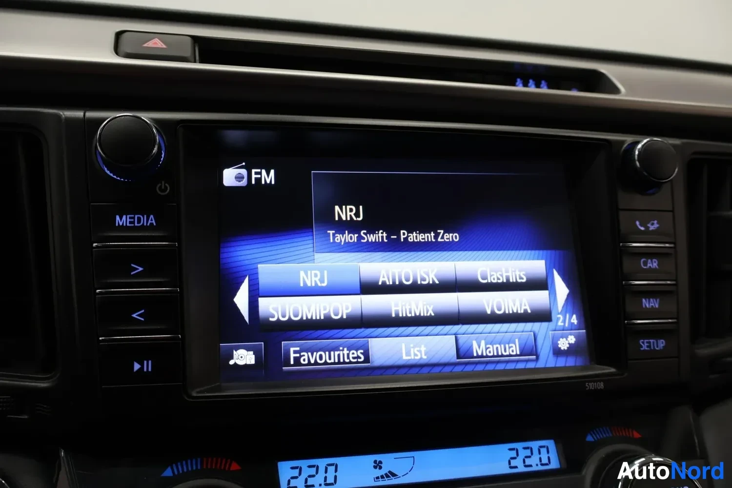 Yksityiskohta: Center console of a car with an infotainment screen showing the radio interface for NRJ by Taylor Swift - Patient Zero. The screen also displays several radio s