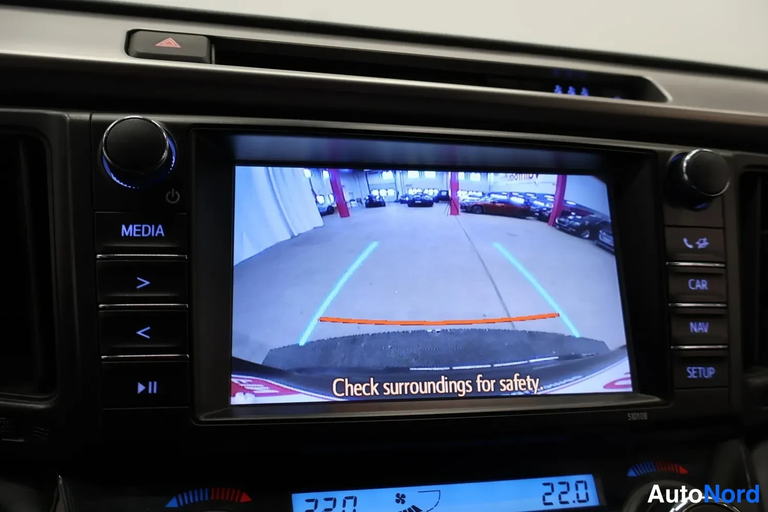 Muu: Kojelaudan infotainment-näyttö näyttää peruutuskameran kuvan ohjausviivoineen ja tekstillä 'Check surroundings for safety.', alapuolella lämpötilanäyttö 22,0.