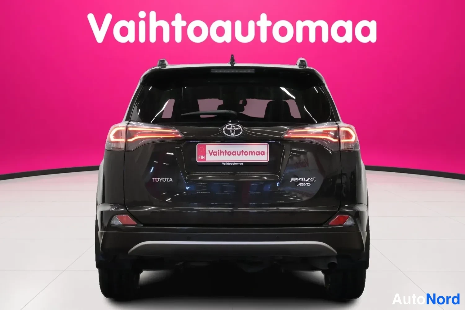 Takaa: Takavaloit, Toyota-logo ja RAV4 AWD -nimike selkeästi näkyvissä mustan auton takana. Taustalla punainen seinä ja Vaihtoautoomaa-tarrat.