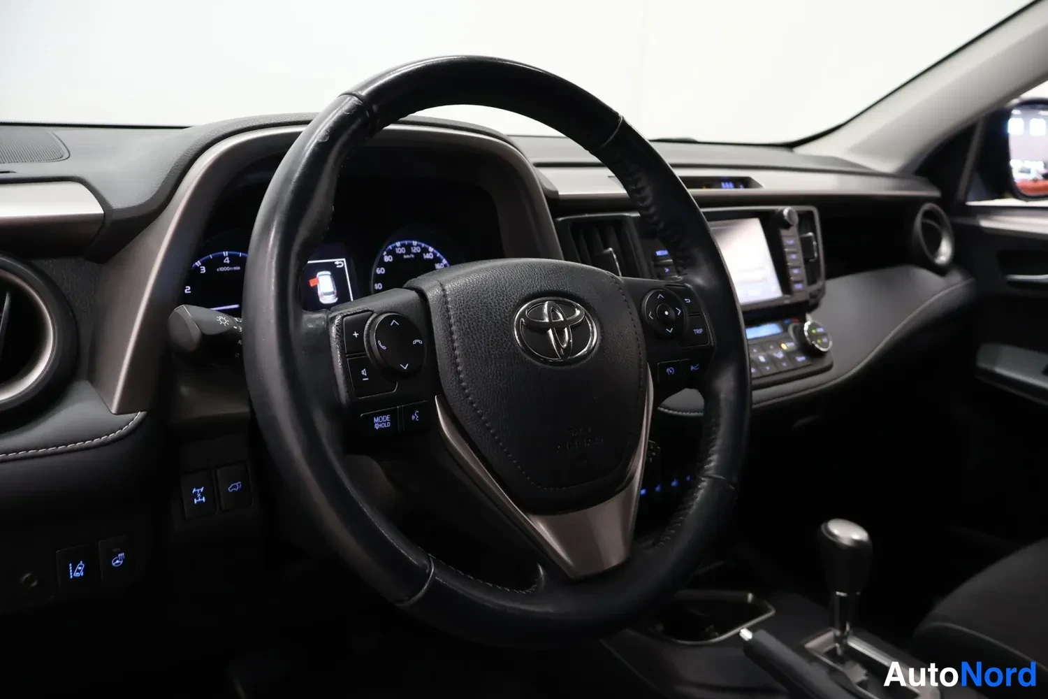 Ohjaamo: Lähikuva ajoneuvon ohjaamosta: musta nahkainen ohjauspyörä Toyota-logolla, keskellä kosketuspaineinen mittaristo ja taustavalaistut painikkeet. Automaattivaihdi