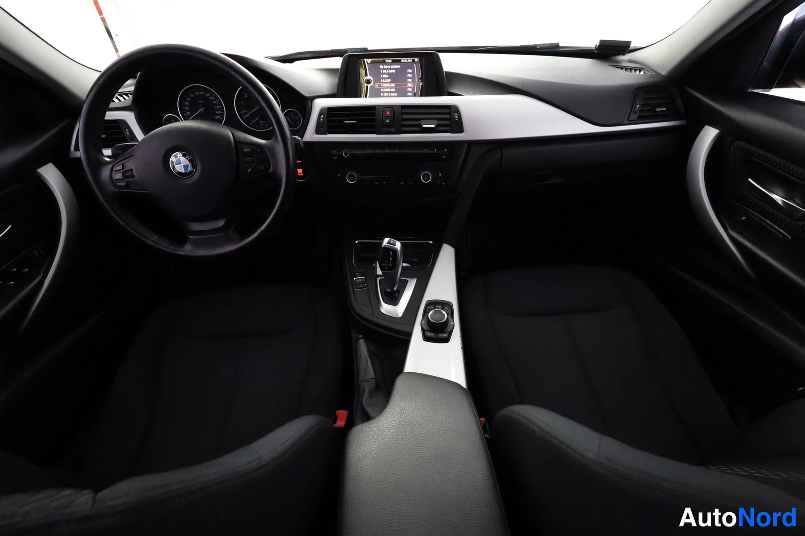 Sivulta: Auton sisätila kuvattuna ohjaamosta taakse päin: BMW-ohjauspyörä, kojetaulu, näyttö jossa radiolistaus, sekä automaattivaihteen valitsin ja iDrive-ohjain keskik