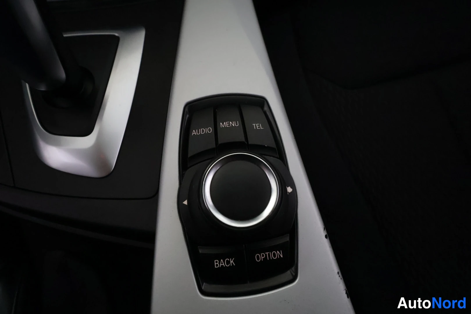 Yksityiskohta: Keskikonsolin iDrive-ohjain lähikuvassa: pyörivä nuppi ja AUDIO-, MENU-, TEL-, BACK- ja OPTION-painikkeet.
