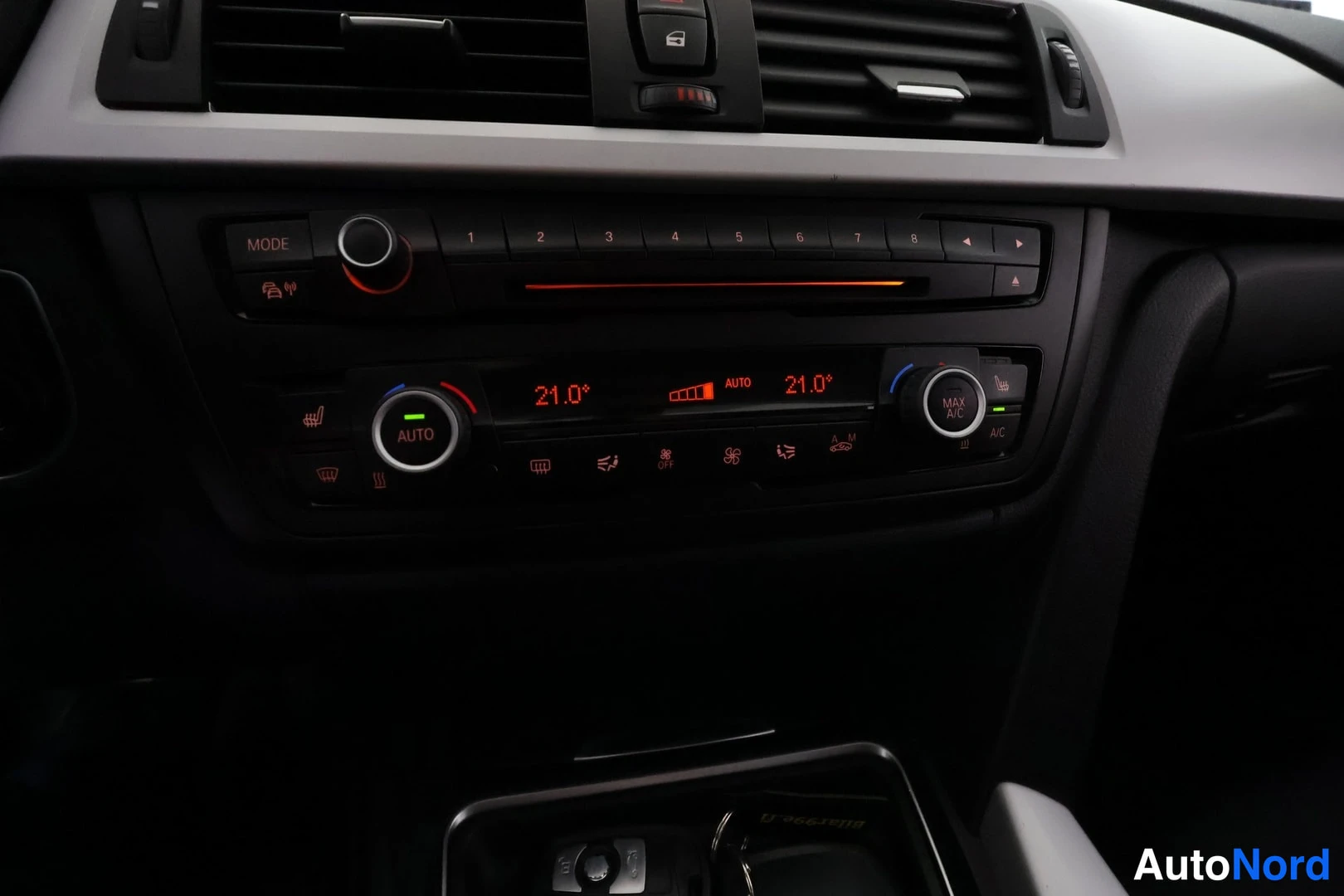 Muu: Kojelaudan keskikonsolin lähikuva: ilmastointipaneelin näytöt näyttävät 21,0° ja AUTO-merkinnän kummallekin puolelle; paneelissa myös radiomuistipaikkojen näppä