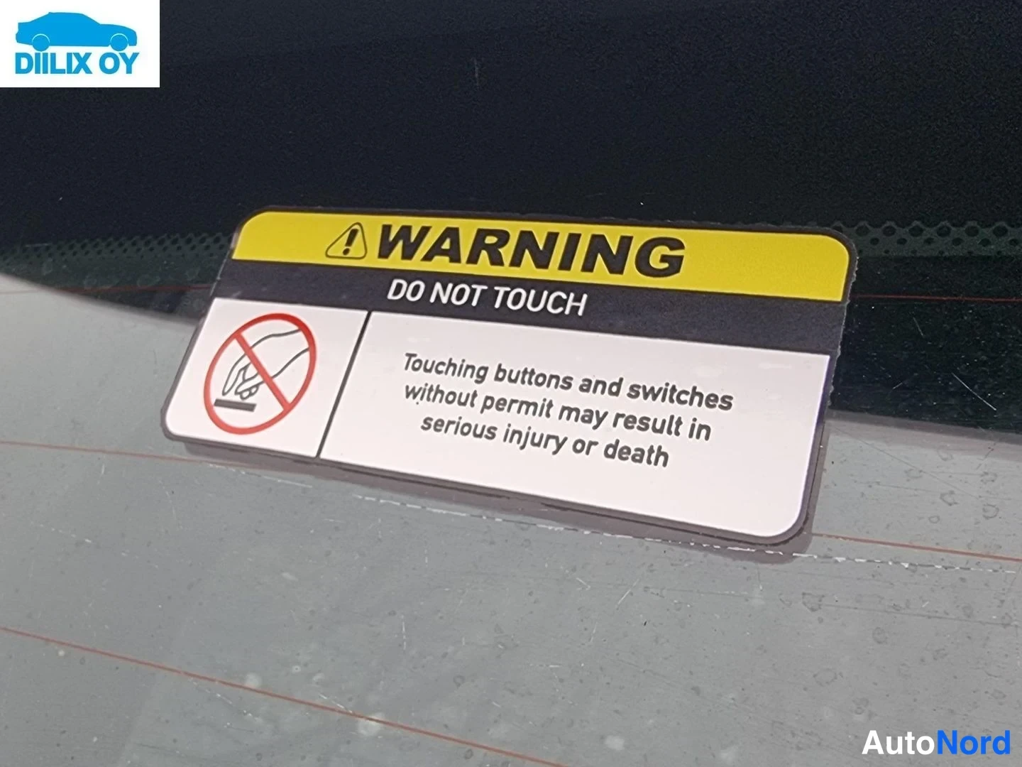 Yksityiskohta: Keltamustea 'WARNING DO NOT TOUCH' -varoitustarra auton tuulilasin tai ikkunan sisäpinnalla, jossa on kielletyn kosketuksen kuvake ja englanninkielinen varoitus