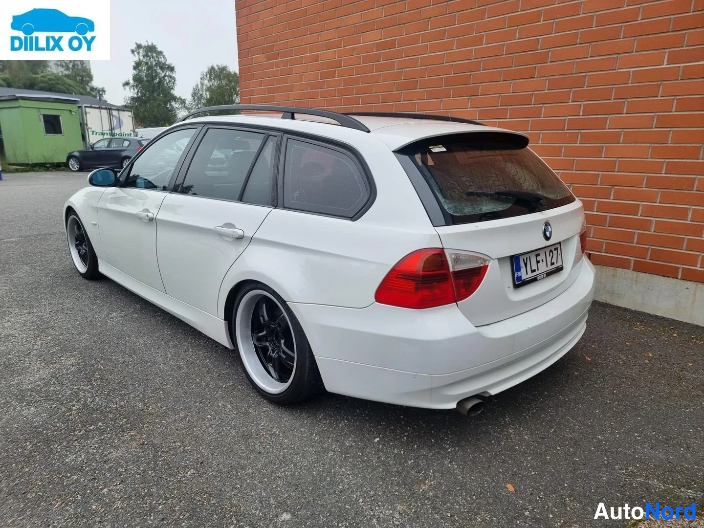 Takaviisto: Valkoinen BMW 3-sarjan farmari kuvattuna takavasemmalta: takavalot, rekisterikilpi, mustat vanteet, kattokaiteet ja takaspoileri näkyvät, taustalla punatiilinen
