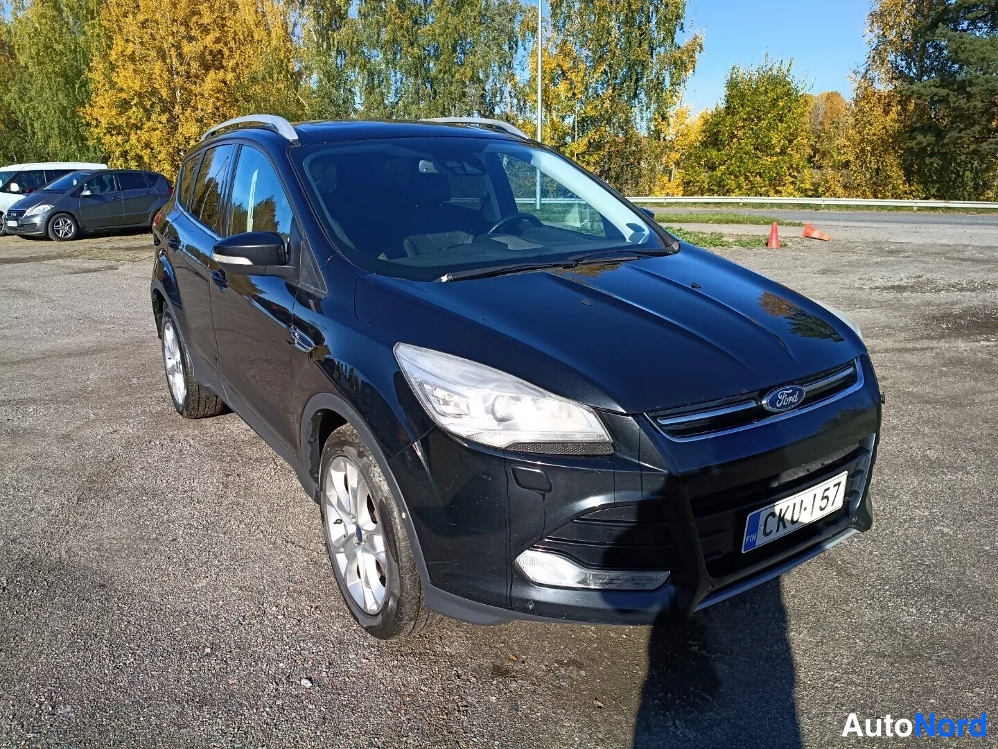 Etuviisto, nokka oikealle: Musta Ford Kuga -henkilöauto vinosta etunäkymästä, etusäleikkö ja Ford-merkki näkyvissä, rekisterikilpi.