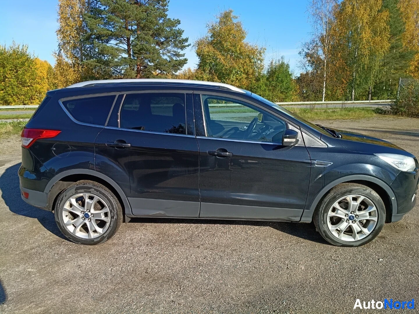 Etuviisto, nokka oikealle: Tumma Ford Kuga -maasturin etukulmakuva, etupaneeli ja vasen kylki näkyvät hiekkaisella parkkipaikalla, kattoharja ja hopeavanteet