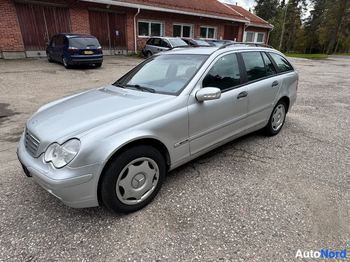 Etuviisto, nokka vasemmalle: Valkohopea farmariliina-auto pysäköitynä asvalttiin parkkipaikalle, auton etupuoli ja vasen kylki näkyvät.