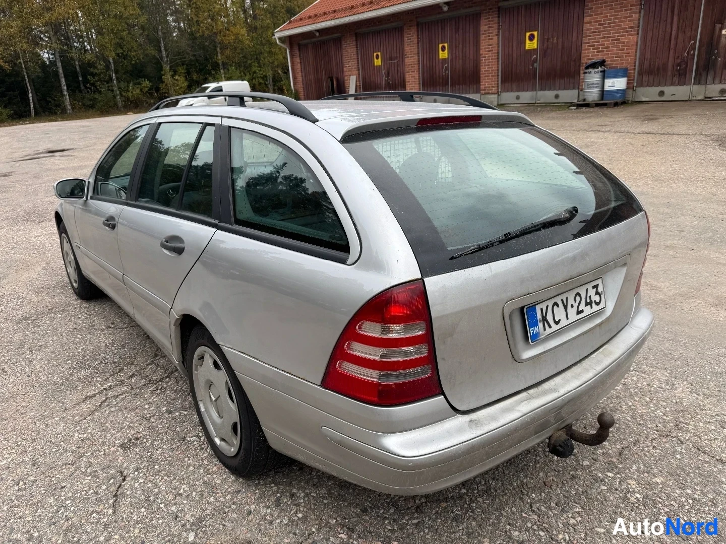 Takaviisto: Mercedes-Benz farmaritakana oikealta takaviistolta, hopeakorinen, kattokaiteet ja vetoaisa näkyvissä, rekisterikilpi.