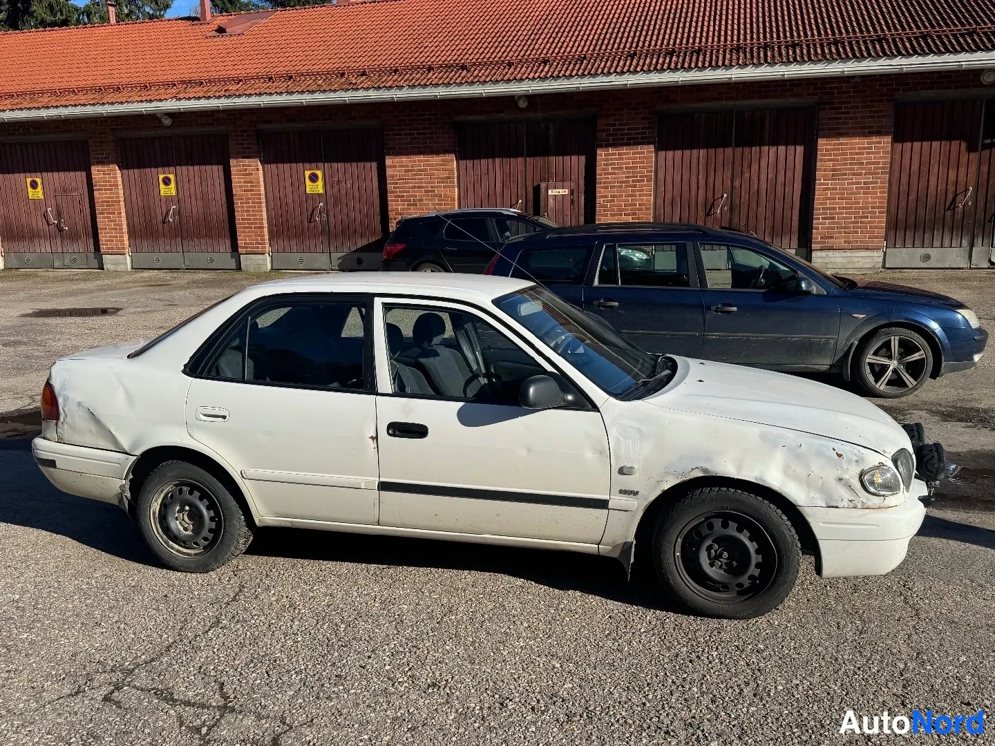 Etuviisto, nokka oikealle: Valkoinen sedan kuvattu sivulta, kori ruhjeilla ja naarmuilla, punatiilikattoinen talli taustalla.
