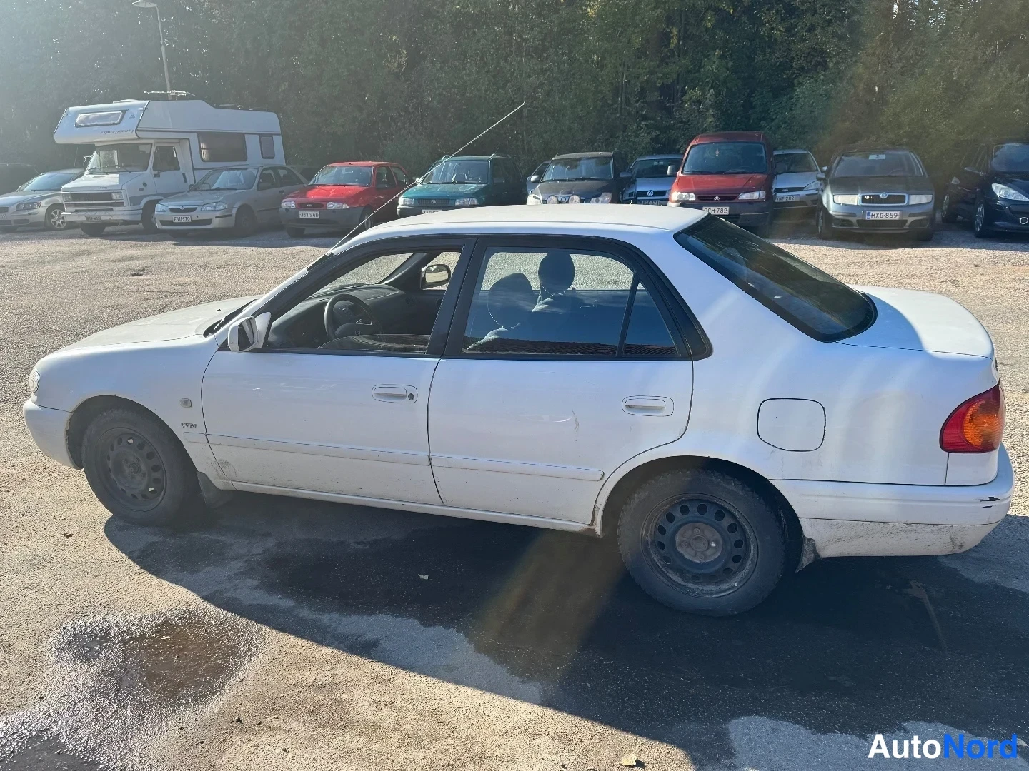 Etuviisto, nokka vasemmalle: Valkoinen Toyota Corolla -sedan vasemmalta viistolta: etuvalo ja keula vasemmassa reunassa, koko vasen kylki ja takavalo näkyvissä.