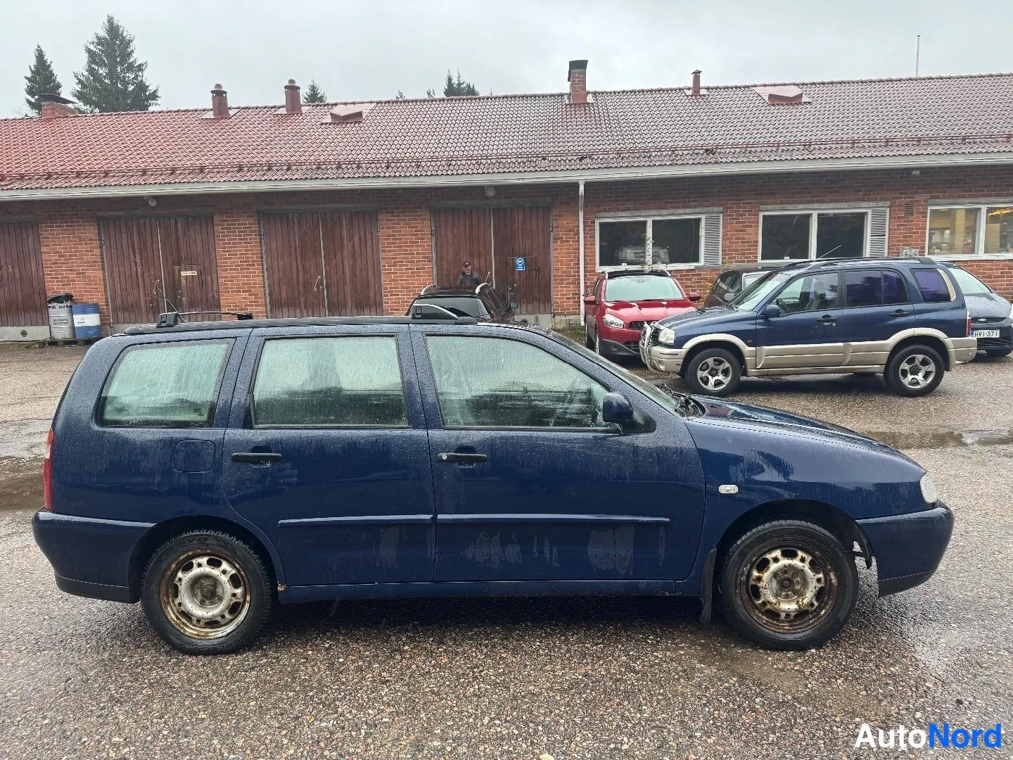 Etuviisto, nokka oikealle: Sininen farmariauto sivulta etuosasta kuvattuna, märkä päällyste ja ruosteiset vanteet.