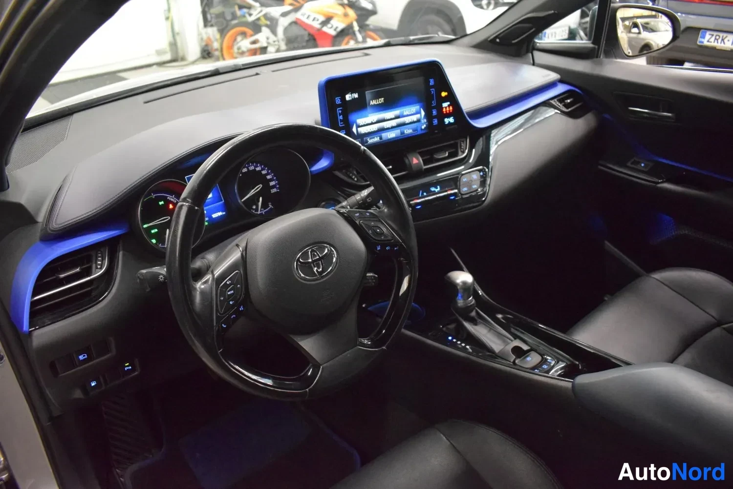 Edestä: Autojen sisätilakuva: ohjauspyörä Toyota-logolla, kojelauta ja kosketusnäytöllinen mediapuhelinjärjestelmä, automaattivaihteiston valitsin sekä mustat nahkapenk