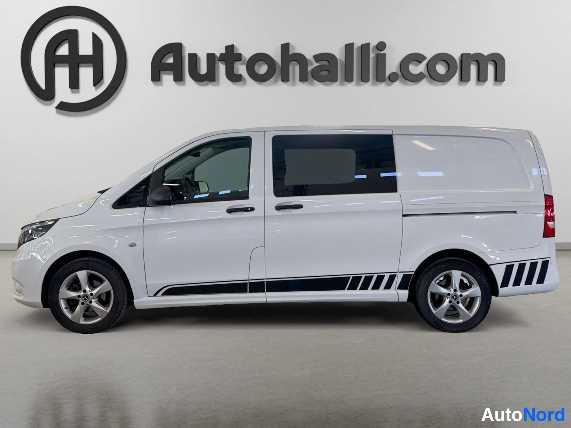 Etuviisto, nokka vasemmalle: Mercedes-Benz Vito -pakettiauto valkoisena, etukolmannesvasen näkymä studioissa, mustaa grafiikkaa ovissa ja alusta.