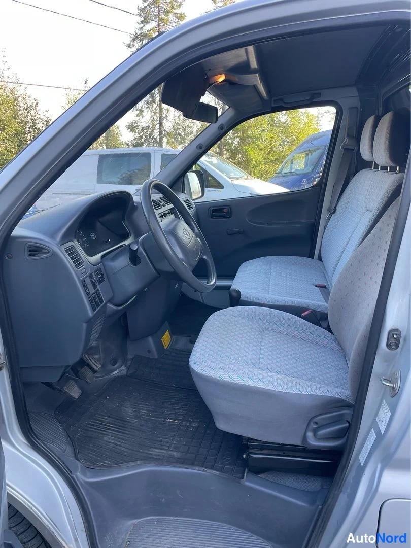Etuistuimet: Driver's cabin interior showing grey fabric bucket seats, dashboard layout, three-spoke steering wheel and floor mats. Manual transmission gear lever is visible