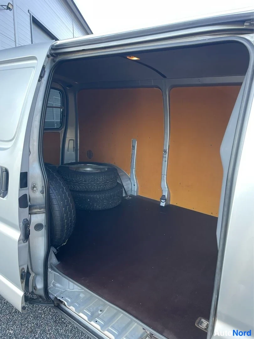 Tavaratila: Rear cargo compartment view through open tailgate showing spare tires stacked on driver's side and wood-finished bulkhead wall.