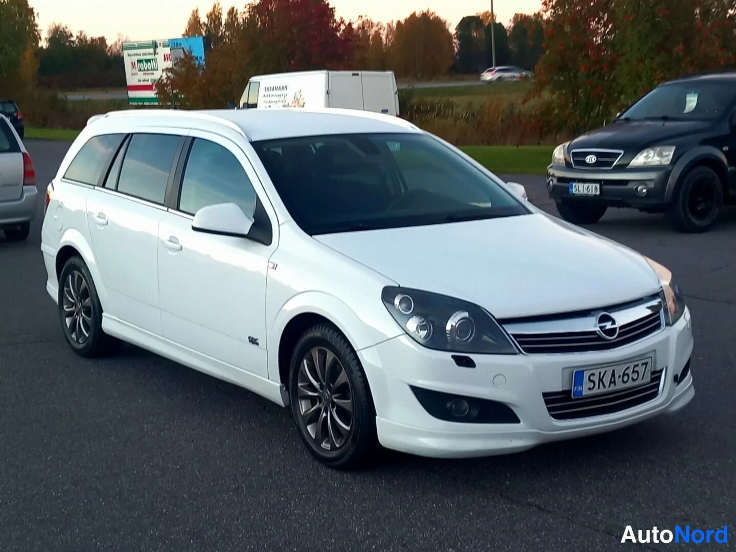 Etuviisto, nokka oikealle: Valkoinen Opel Astra -farmariauto etukulmasta vasemmalta, rekisteritunnus, tummat kevytmetallivanteet.