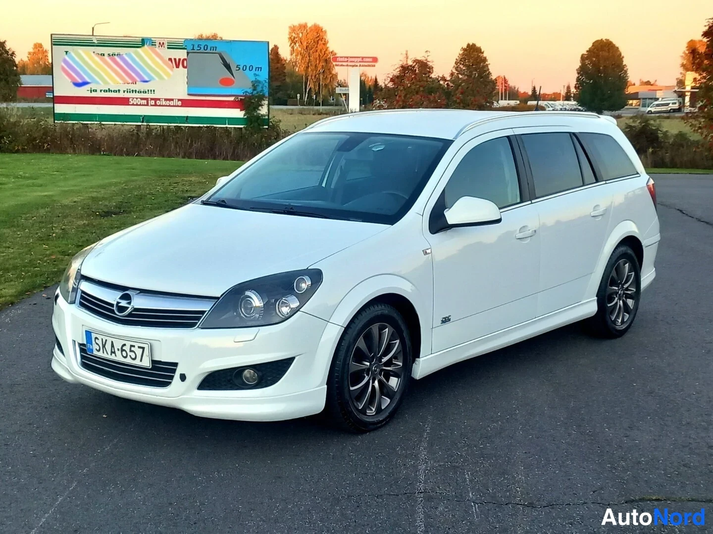 Etuviisto, nokka vasemmalle: Valkoinen Opel Astra -farmariauto etukulmasta, rekisteritunnus, tummat kevytmetallivanteet.