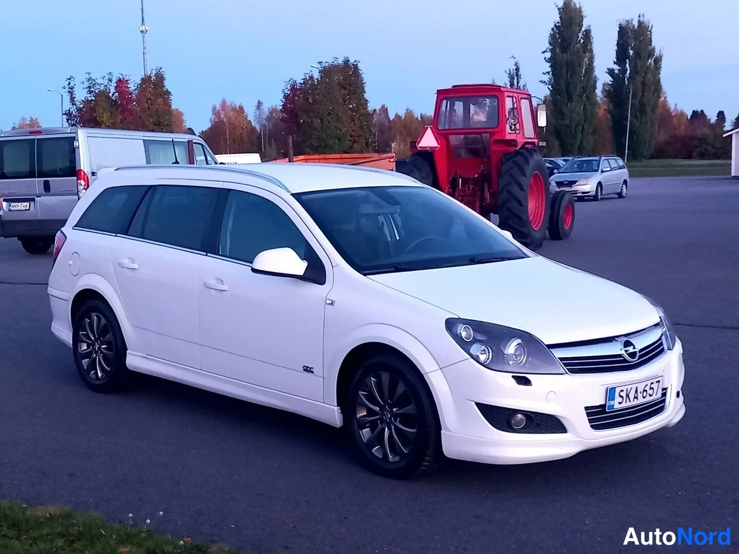 Etuviisto, nokka oikealle: Valkoinen Opel Astra -farmariauto etukulmasta vasemmalta, rekisteritunnus, tummat kevytmetallivanteet, taustalla punainen traktori.