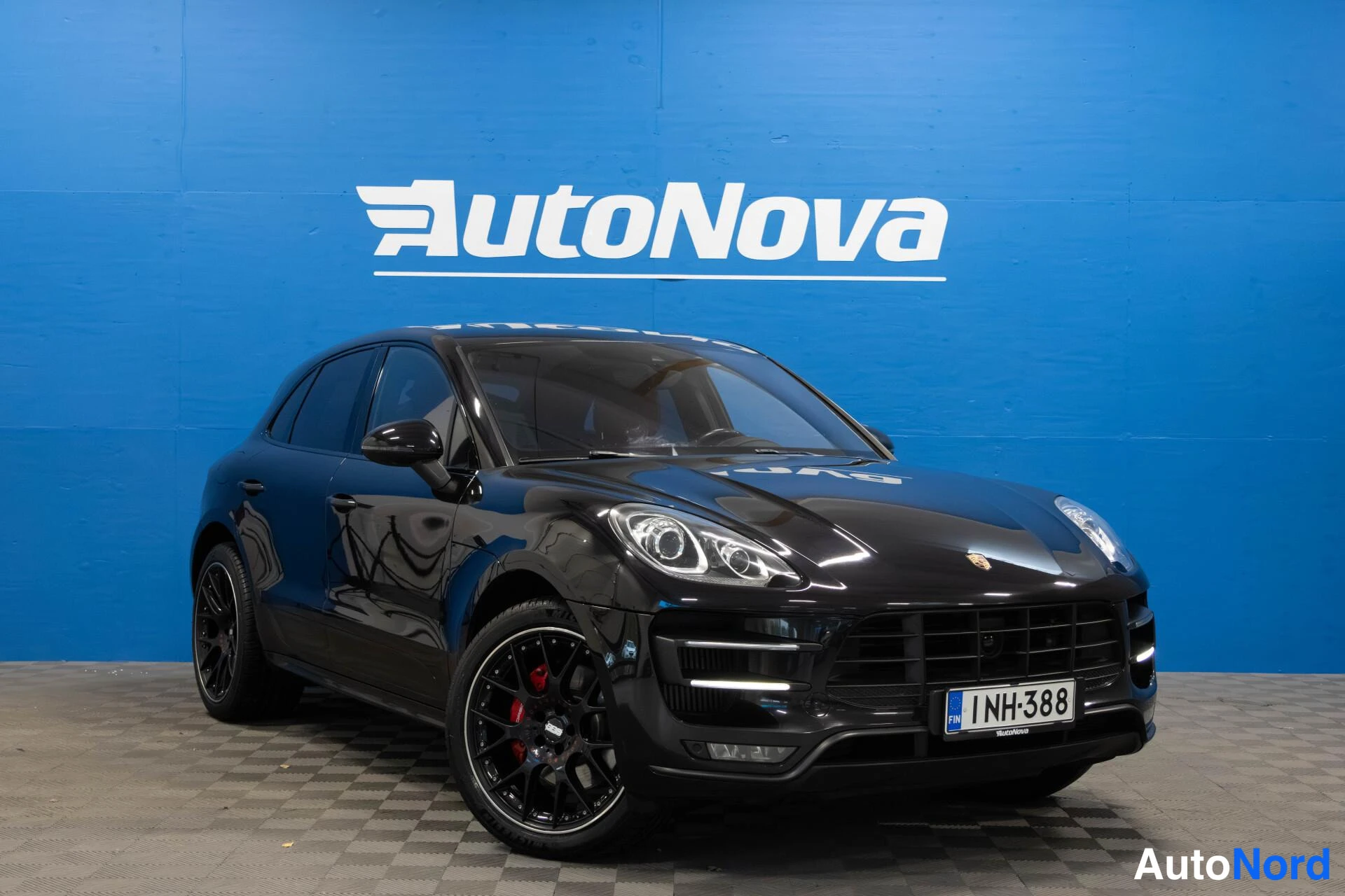 Etuviisto, nokka oikealle: Mustan Porsche Macan -maasturin etukulmanäyttö, rekisteritunnus, sininen AutoNova-taustaseinä.
