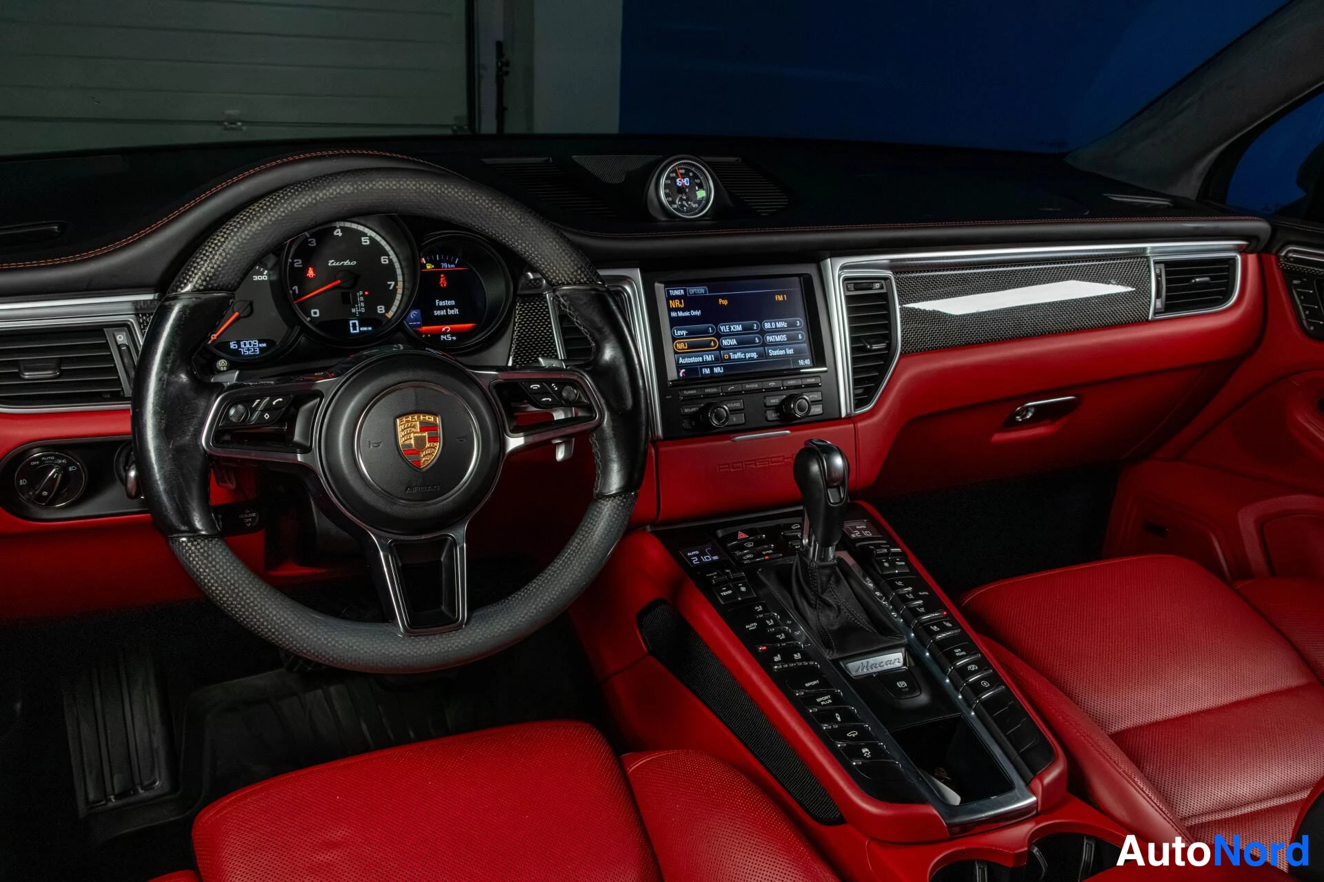 Yksityiskohta: Ohjaamo ylhäältä: Porsche-ohjauspyörä, punaiset istuimet ja panelit, keskusnäyttö radio-ohjelmalla, vaihteenvalitsin ja konsolin painikkeet.
