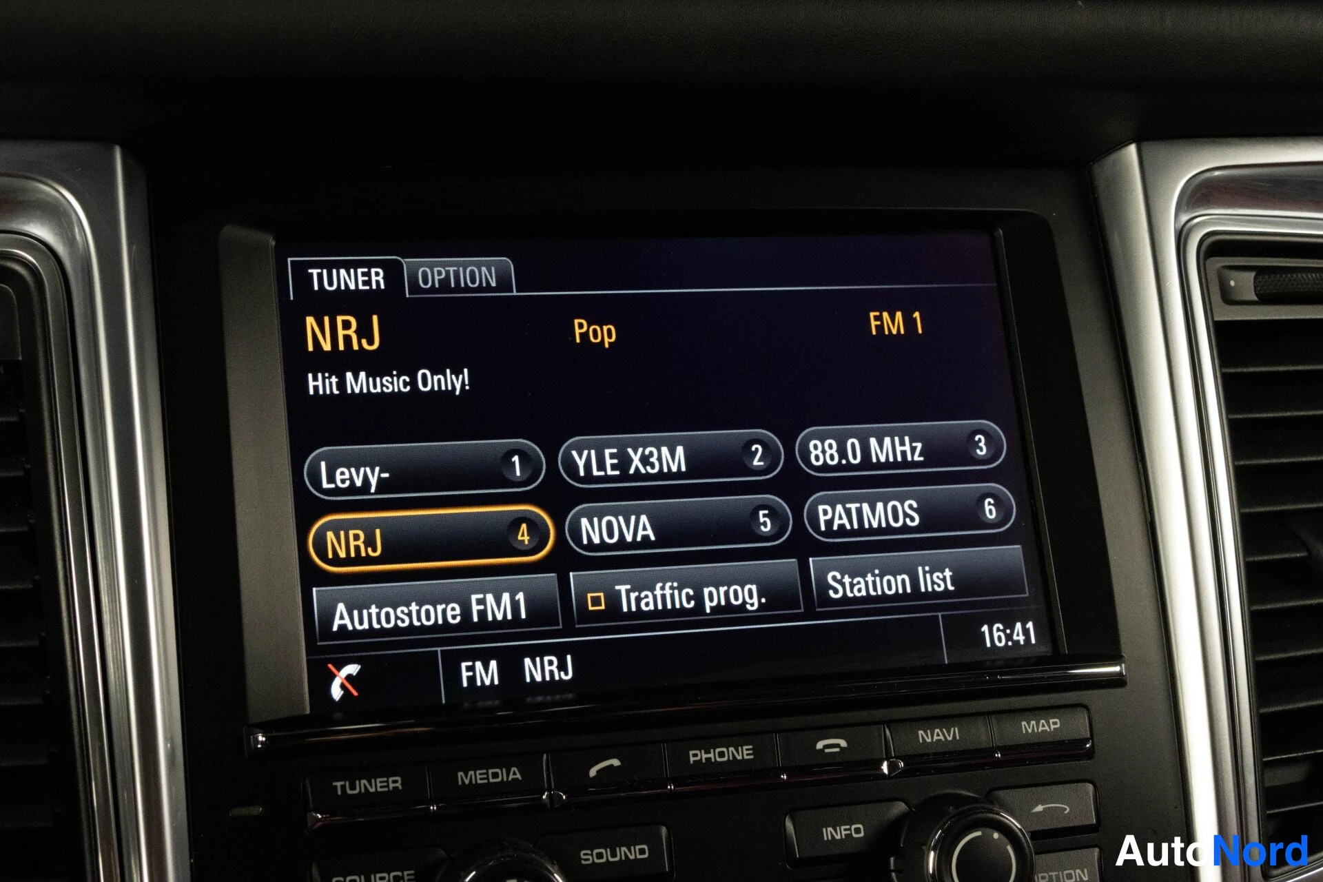 Yksityiskohta: Autoradion näyttö: TUNER-option FM 1 -kanava NRJ, esiasetettuina YLE X3M, NOVA ja PATMOS, kello 16:41.