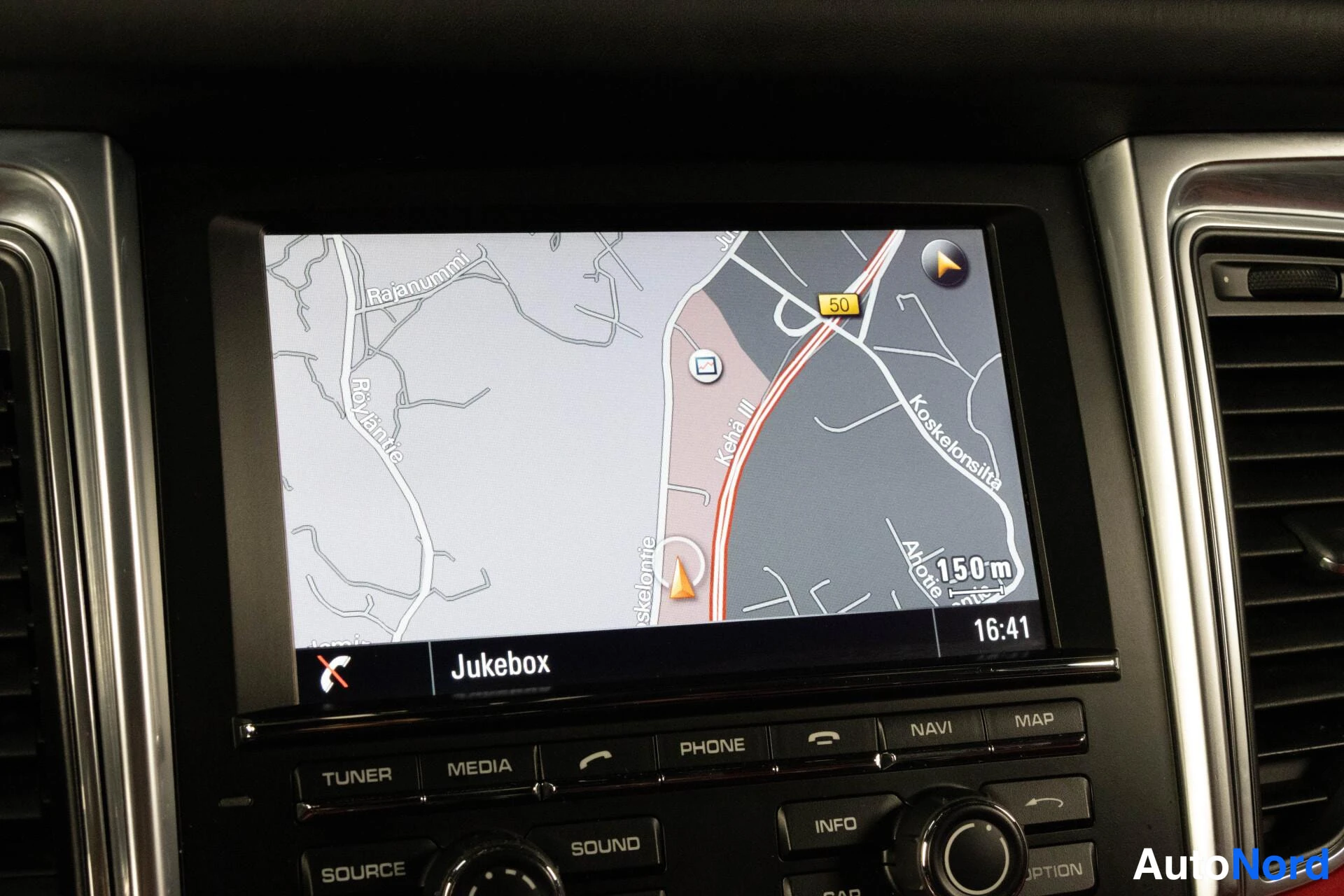 Yksityiskohta: Auton navigaatiolaitteen näyttö karttanäkymässä paikannimillä Rajamumm, Koskelon itsa ja Kehä III, kellonaika 16:41.