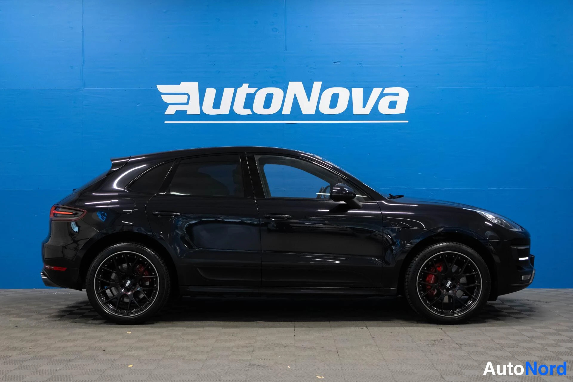 Sivulta: Mustan Porsche Macan -maasturin sivunäyttö vasemmalta puolen, mustat vanteet ja punaiset jarrusatulat, sininen AutoNova-taustaseinä.