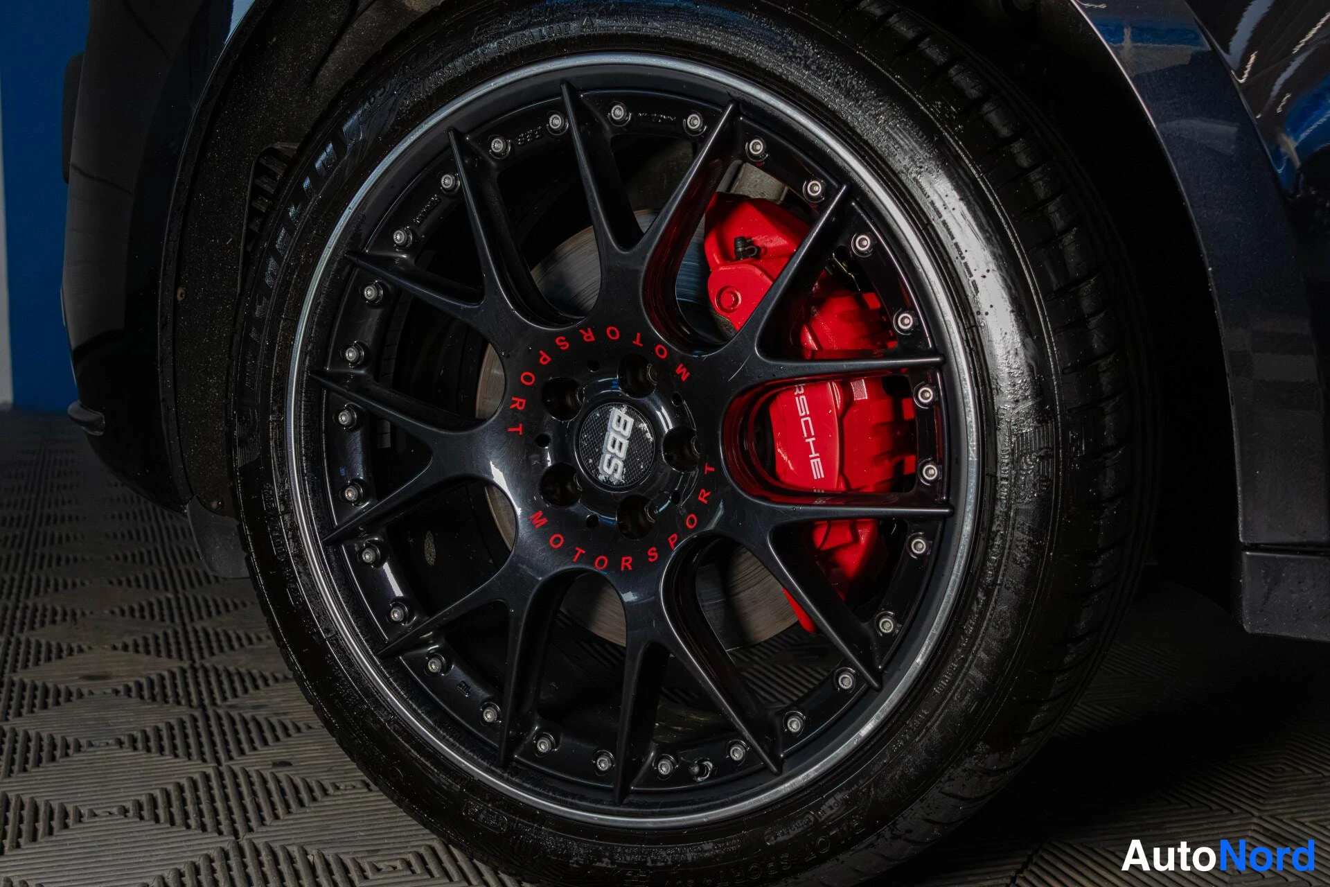 Yksityiskohta: Close-up of a black multi-spoke BBS Motorsport alloy wheel with red Porsche brake calipers visible behind it. Tires show some wear. Shot is taken from a low ang