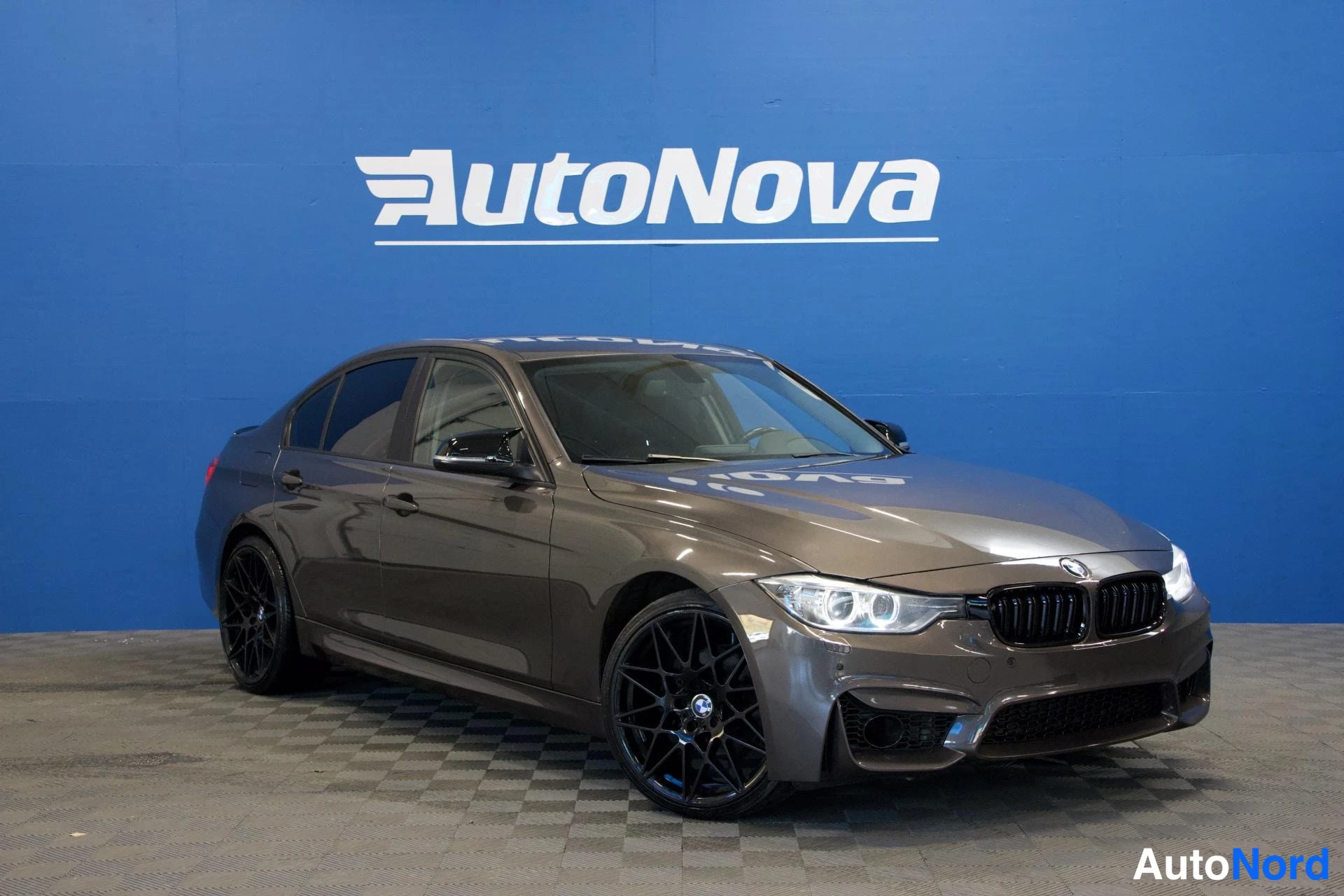 Etuviisto, nokka oikealle: BMW 320 sedan etukulmasta oikealta, tummanruskea, mustat kevytmetallivanteet, sininen AutoNova-taustaseinä.