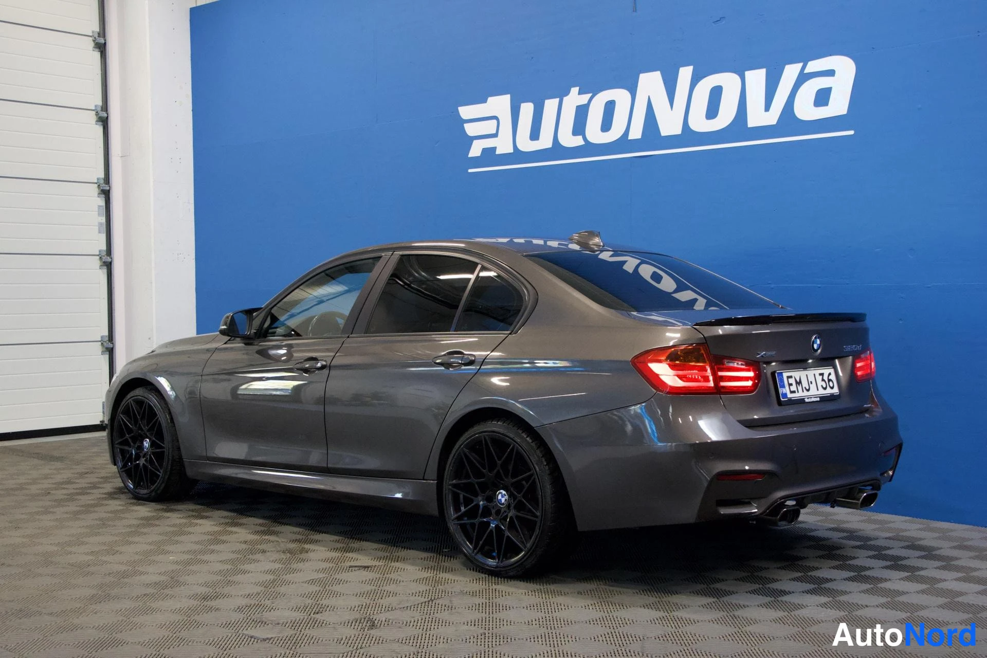 Takaviisto: BMW-sedan takakulman sivulta, tummanharmaa runko, mustat kevytmetallivanteet, neljä pakoputkea, rekisteri, sininen AutoNova-tausta.