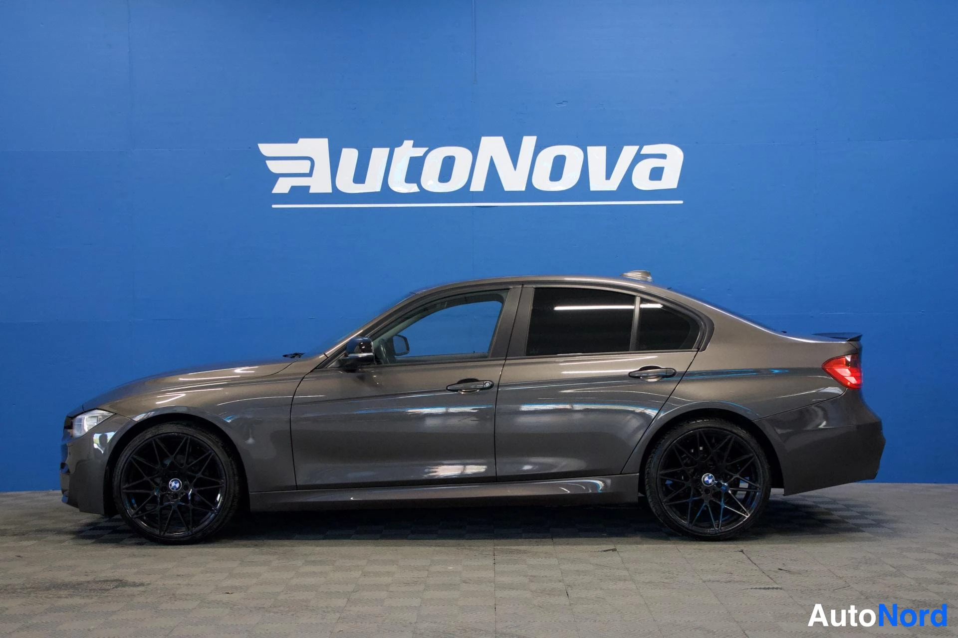 Etuviisto, nokka vasemmalle: BMW-sedan sivuprofiili vasemmasta etukulmasta, tummanharmaa runko, mustat vanteet, sininen AutoNova-tausta.