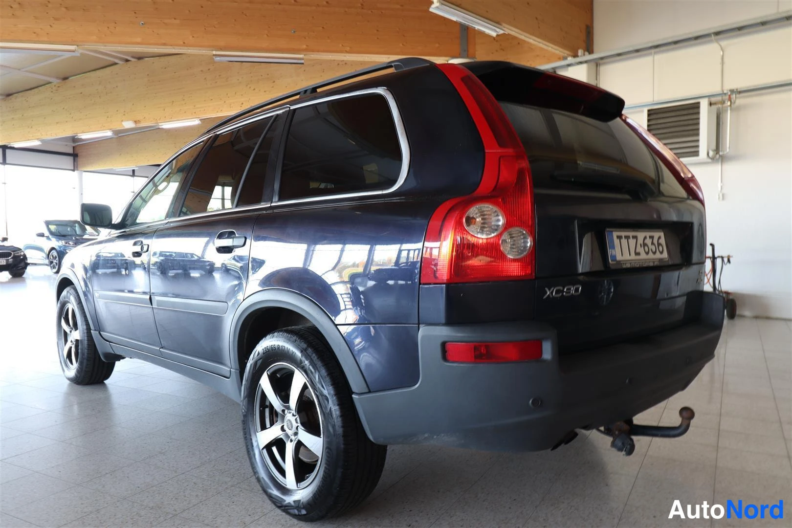 Takaviisto: Tummansinisen Volvo XC90:n takakulma sisätiloissa; takavalot, XC90-merkintä, rekisterikilpi ja takana vetokoukku.