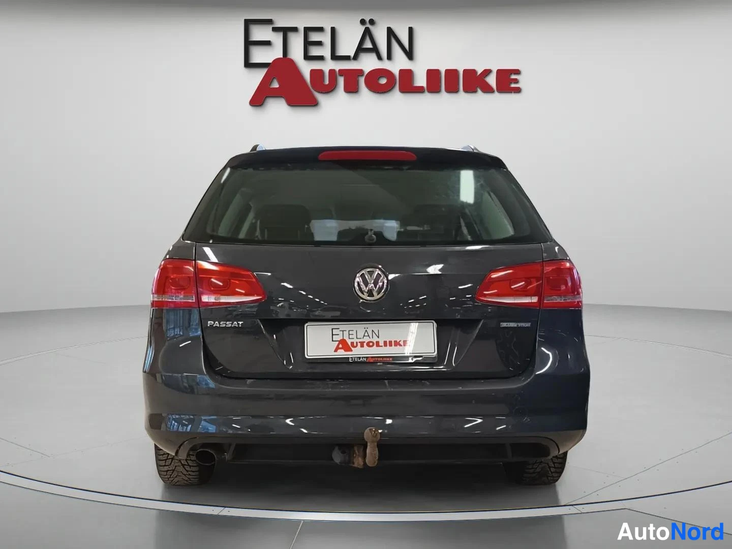 Takaa: Suoraan takaa kuvattu tummanharmaa Volkswagen Passat -farmari, jossa on takaosaan kiinnitetty kytkinkuula ja vara- merkitys rekisterikilven paikalla.