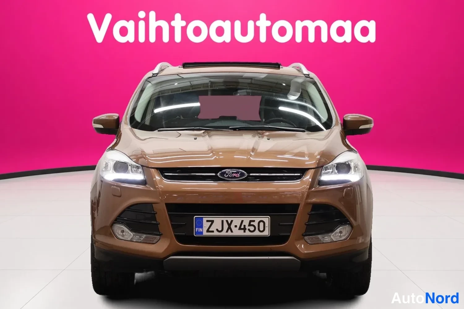 Edestä: Ruskea Ford Kuga edestäpäin kuvattuna,Ford-merkki jäähdyttimen säleikössä ja rekisteritunnus.