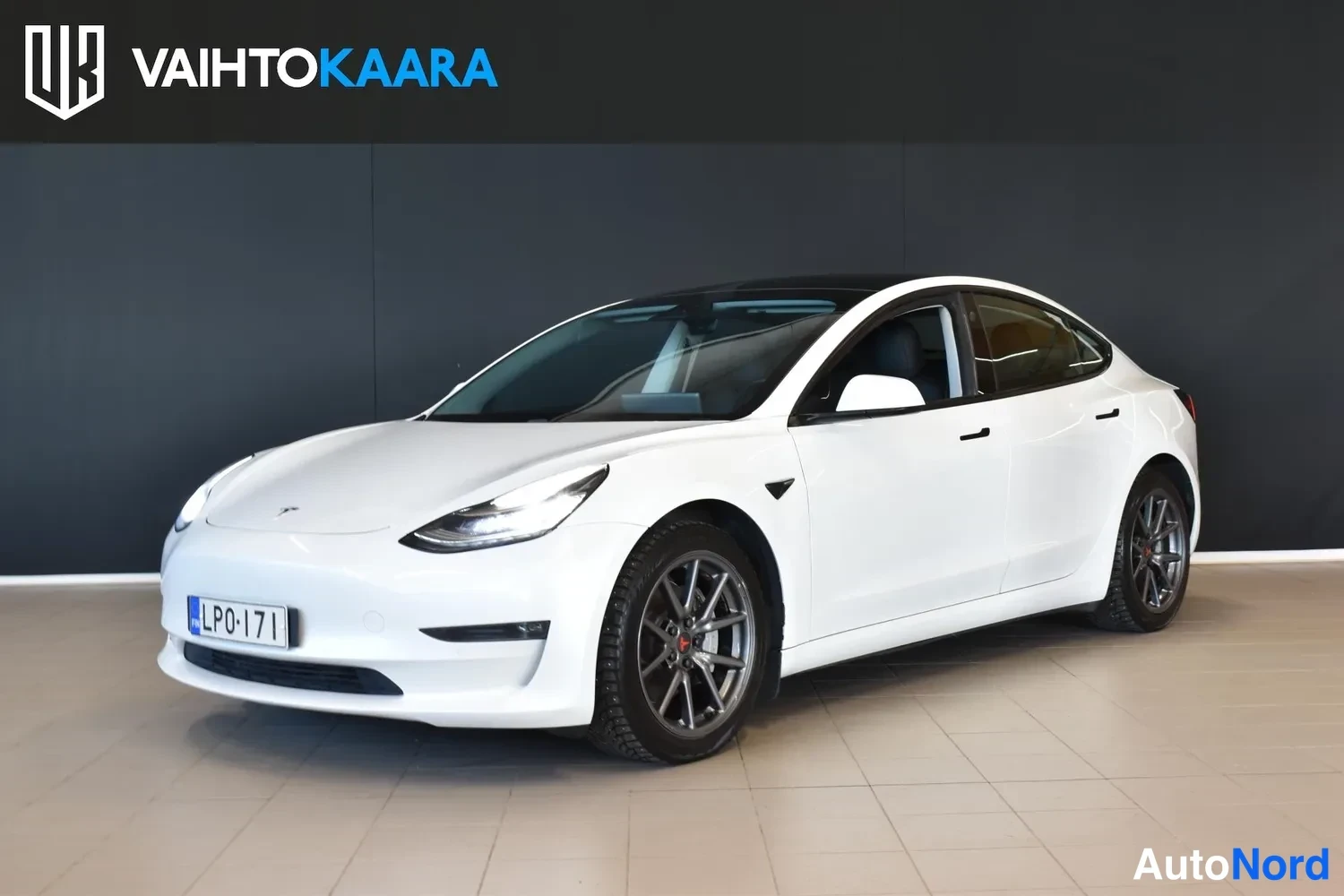 Etuviisto, nokka vasemmalle: Tesla Model 3 -sedanin etukulma showroomissa; valkoinen koripaneli, tumma lasikatto, harmaat kevytmetallivanteet ja rekisterikilpi
