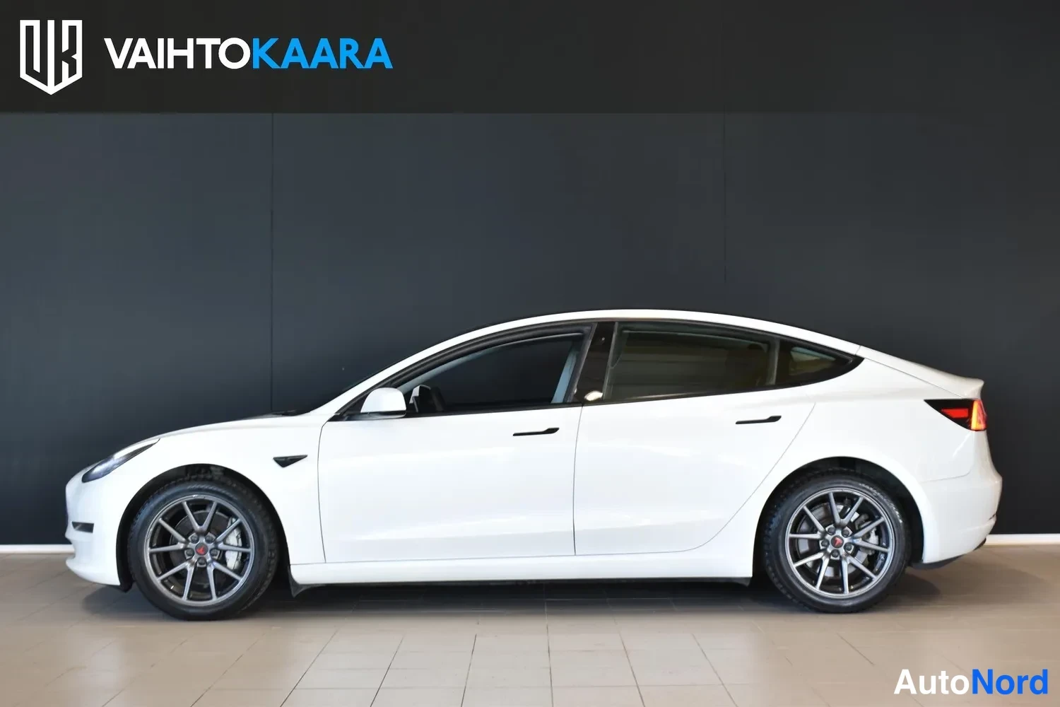 Sivulta: Tesla Model 3 -sedanin sivuprofiili showroomissa; valkoinen koripaneli, tumma lasikatto ja harmaat kevytmetallivanteet