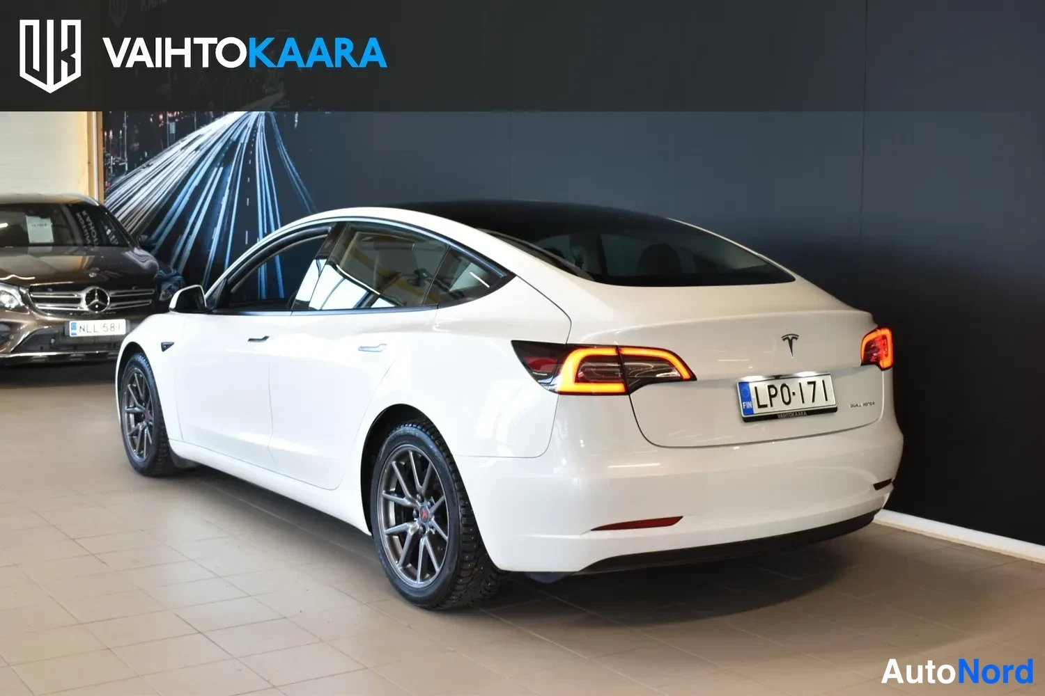 Takaviisto: Tesla Model 3 -sedanin takakulma showroomissa; valkoinen kori, syttyneet takavalot, tumma lasikatto ja rekisterikilpi