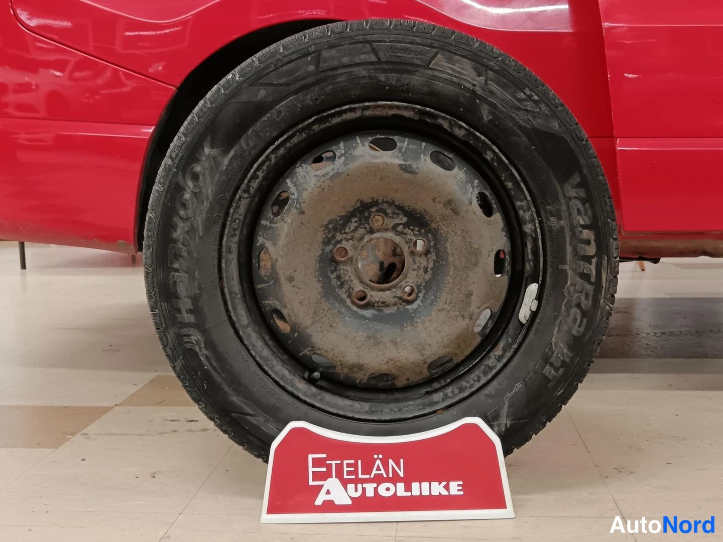 Yksityiskohta: Toinen pyörä lähikuvassa: Halkeillut Hankook Ventra -renkaan sivuseinä ja roosteventtiili, punainen Etelän Autoliike -kilpi lattialla.