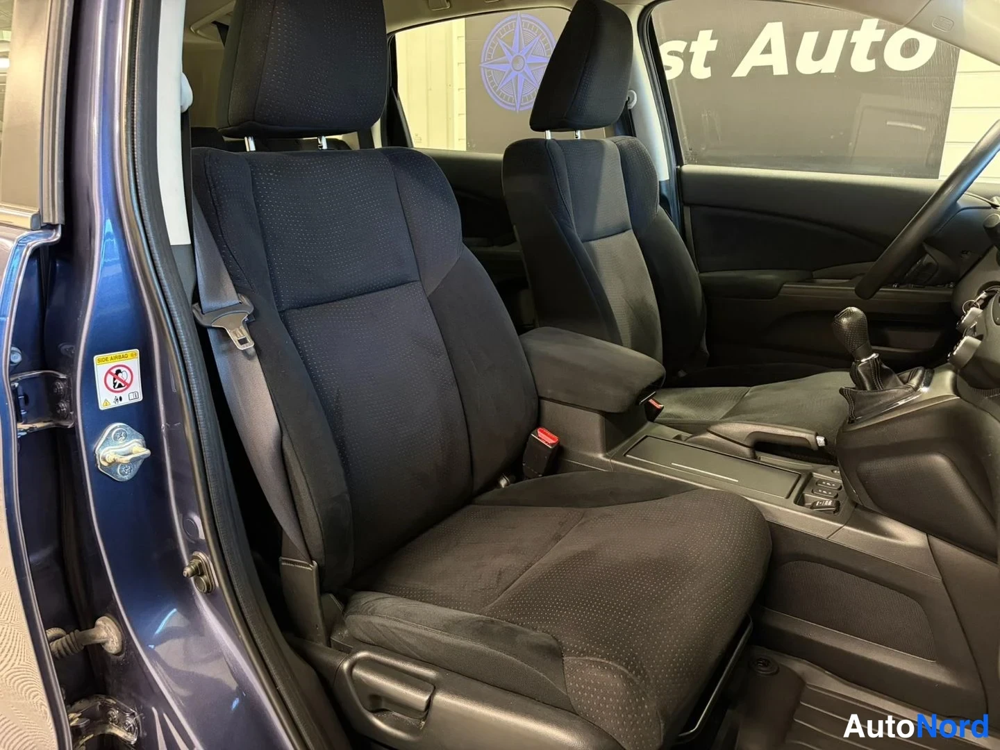 Muu: Sisätilakuva: Honda CR-V:n etummat istuimet ja matkustamotila, tummat kankaat istuimet, vaihdekeppi ja lattiamatot näkyvät avoimesta kuljettajan ovesta kuvattun