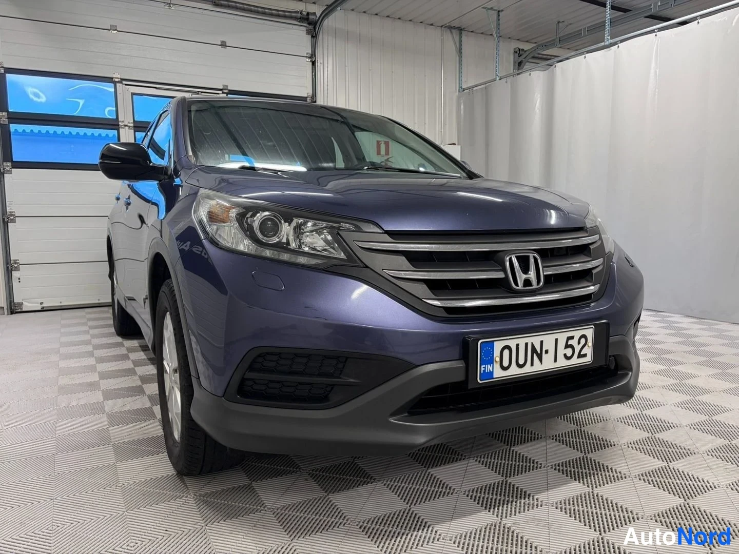 Etuviisto, nokka oikealle: Honda CR-V:n etukulma kuvattuna oikealta edestä autotallissa, tummansininen kori ja rekisterikilpi näkyvät.