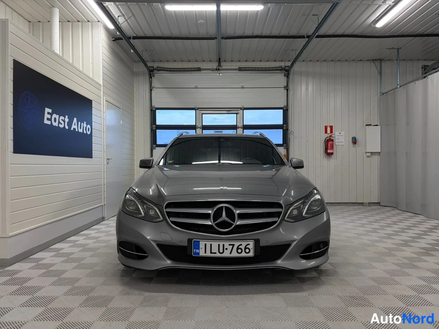 Edestä: Mercedes-Benz E-farmari suoraan edestä kuvattuna autotallissa, harmaan metallinhohtoinen kori, Mercedes-tähti etusäleikössä, rekisteritunnus.