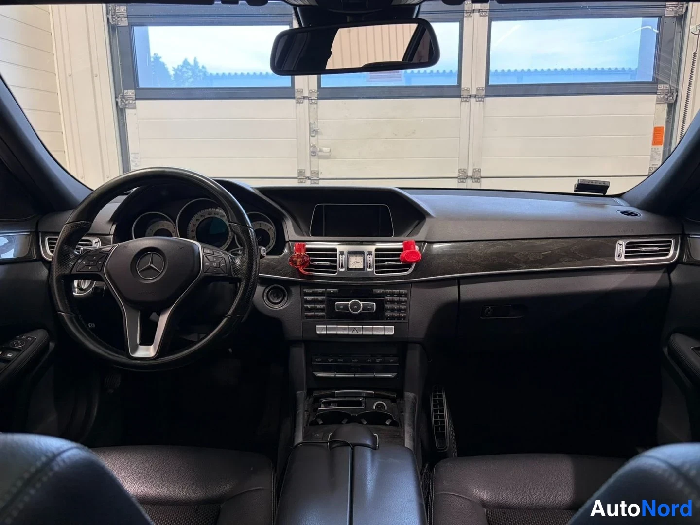 Edestä: Mercedes-Benzin ohjaamo etuosasta kuvattuna: Mercedes-merkitty ohjauspyörä, kojelauta ja keskikonsoli, tumma sisusta.