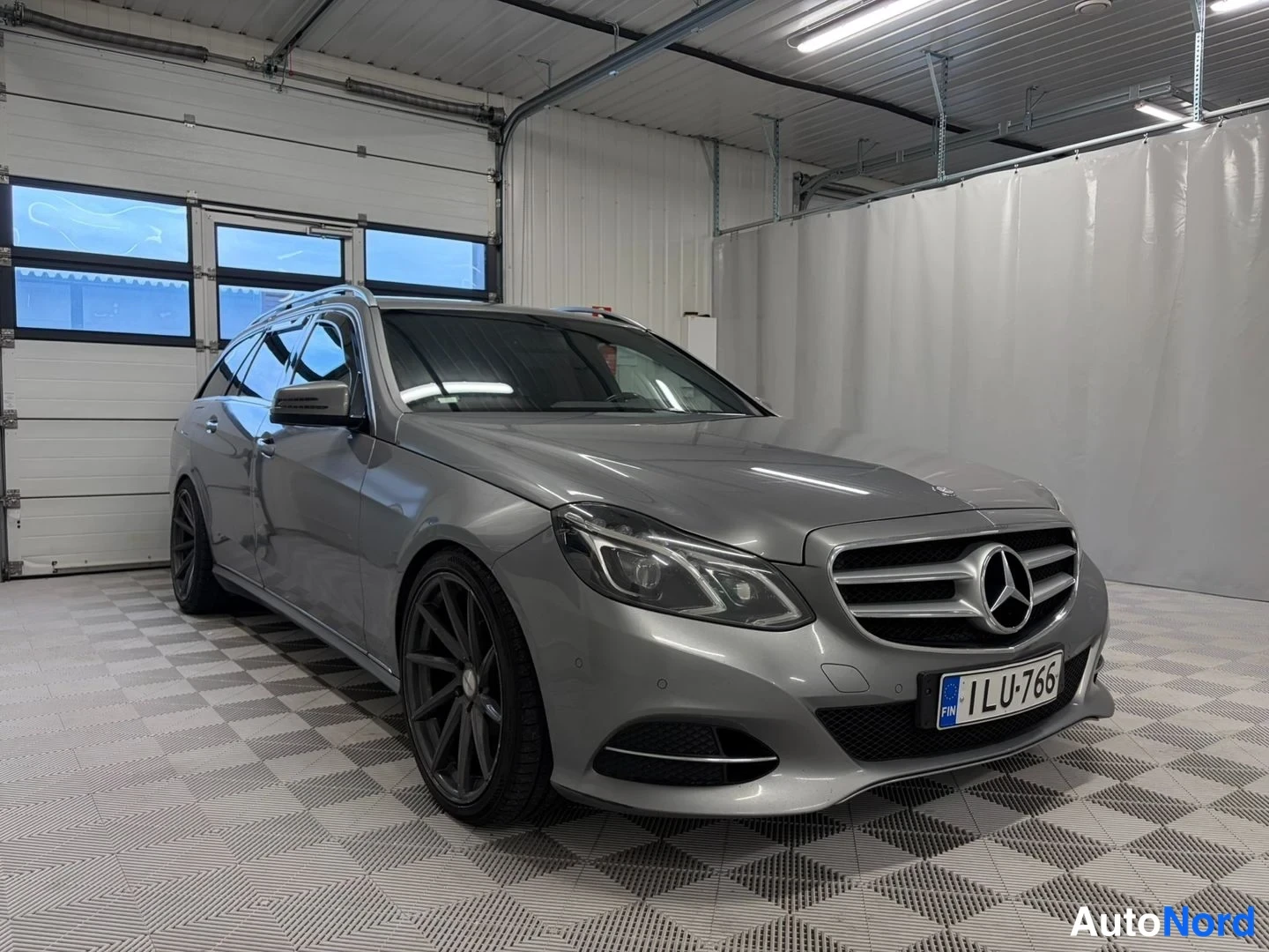 Etuviisto, nokka oikealle: Mercedes-Benz E-farmari sivulta-edestä oikealle kuvattuna sisätilassa, harmaa kori, tummat vanteet, rekisteritunnus.