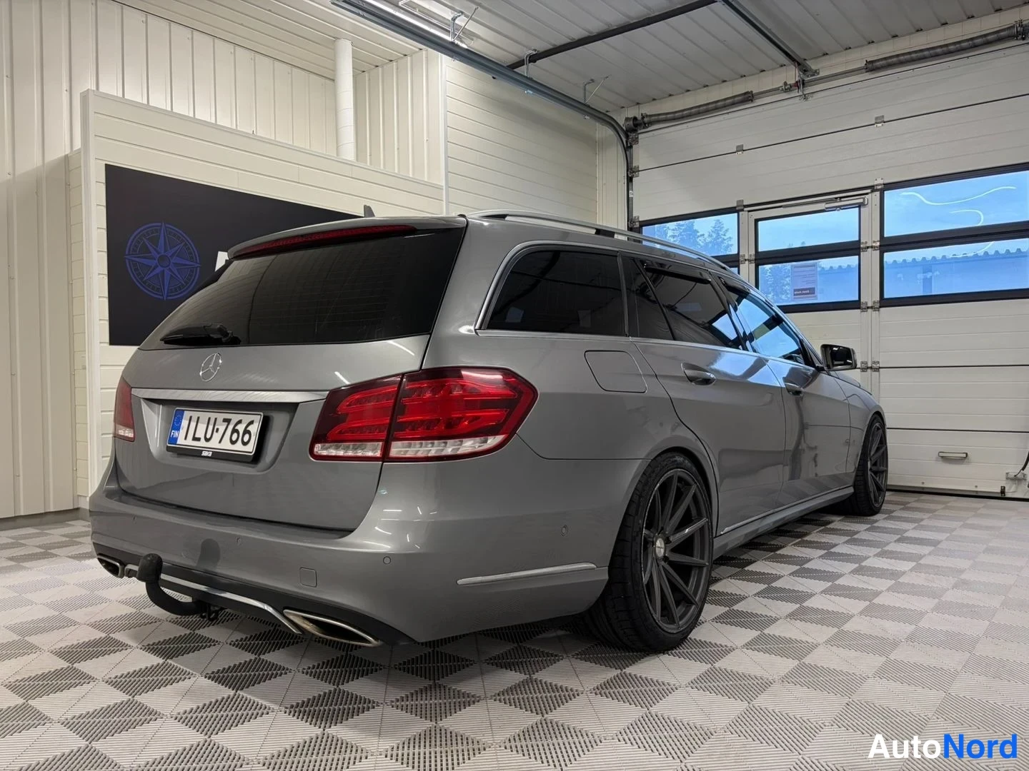 Takaviisto: Mercedes-Benz E-farmari sivulta-takaa vasemmalle kuvattuna autotallissa, harmaan metallinhohtoinen kori, punaiset takavalot, rekisteritunnus.